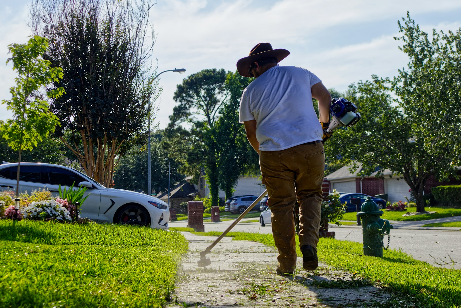 Yard worker trimming weeds along a sunny residential sidewalk